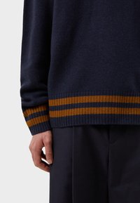 Marinblått stickat tröja med ribbad nederkant som har två horisontella ränder i senapsgul och mörkblå. Närbild av ärm och hand.