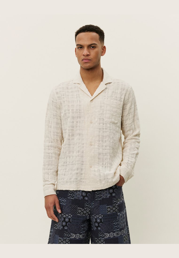 Helles beige Leinenhemd mit einem strukturierten Muster, langen Ärmeln und einem eingeschnittenen Kragen, kombiniert mit marineblauen gemusterten Shorts.