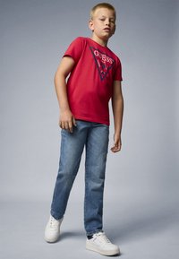 Rød bomull T-skjorte med et trekantet "GUESS" logo design, kombinert med blå denimjeans og hvite atletiske joggesko. Glatt tekstur, avslappet passform.
