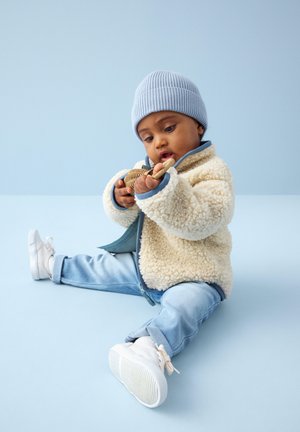 Małe dziecko w niebieskiej czapce, kremowej polarowej kurtce i dżinsach siedzi na podłodze, uważnie trzymając i badając drewnianą zabawkę.