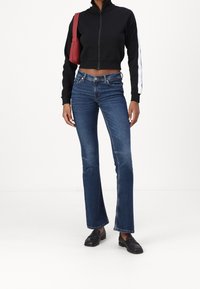 Schwarze, kurz geschnittene Reißverschlussjacke mit weißen Seitenstreifen, kombiniert mit blauen, ausgestellten Jeans und schwarzen Loafers. Roter Handtaschenakzent in der Nähe des Arms.