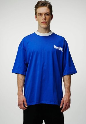 Rövid hajú fiatal férfi laza kék pólót visel, melyen fehér "Reality" felirat található a mellkasán, és egyszínű világos háttér előtt áll.