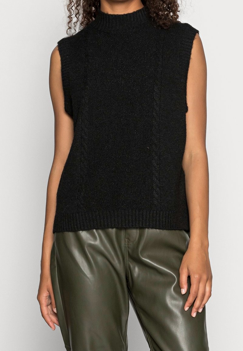 Maglione senza maniche nero con collo alto, caratterizzato da un tessuto lavorato e un orlo a coste. Indossato con pantaloni in pelle verde oliva.