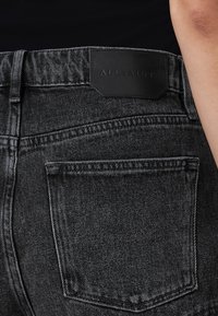 Jeans in denim nero dal design elegante, con etichetta in pelle recante la scritta "ALLSAINTS" e dettagli di cucitura discreti sulla tasca posteriore.