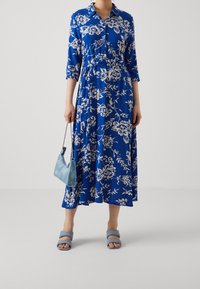 Blomsterkjole i blå med button-down krave, trekvartærmer, indsnævret talje og svajende skørt; parret med en lyseblå håndtaske og sandaler.