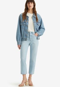 Chaqueta de mezclilla azul claro con un corte relajado, llevada sobre una camiseta blanca y combinada con unos vaqueros azules claros de cintura alta y sandalias de tacón cuadrado beige.