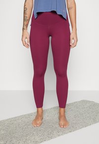 Burgundské legíny s hladkým povrchem a vysokým pasem, doplněné volným modrým sportovním topem. Nohy jsou naboso na šedém povrchu.