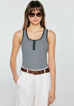 Kobieta w czarno-białym, bezrękawnym topie w paski, białych spodniach z wysokim stanem, brązowym pasku, okrągłych okularach przeciwsłonecznych oraz kolczykach kołach, pozująca z rękami w kieszeniach.