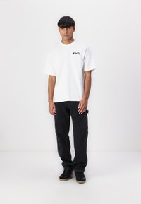 White short-sleeve t-shirt with black logo on chest, paired with black cargo pants and black sneakers, standing against a plain background.