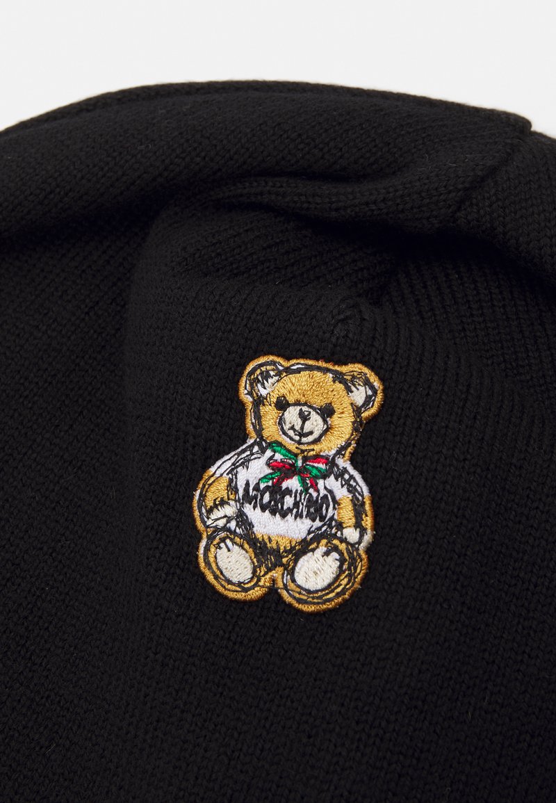 Berretto in maglia nera con patch ricamato di un orso giallo e bianco, che indossa un nastro verde e rosso, con la scritta "Mochino".