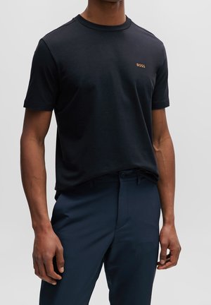 Homme portant un t-shirt noir à manches courtes avec un petit logo doré "BOSS" et un pantalon bleu marine foncé sur un fond clair uni.