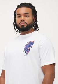 Mand med skæg og dreadlocks iført en hvid T-shirt med et vertikalt grafisk motiv af tre blå og orange kranier med lille tekst nedenfor.