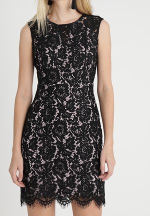 Ærmeløs sort blondekjole med blomsterprint over nude foring, tætsiddende talje og bølget kant, båret af en stående kvinde.