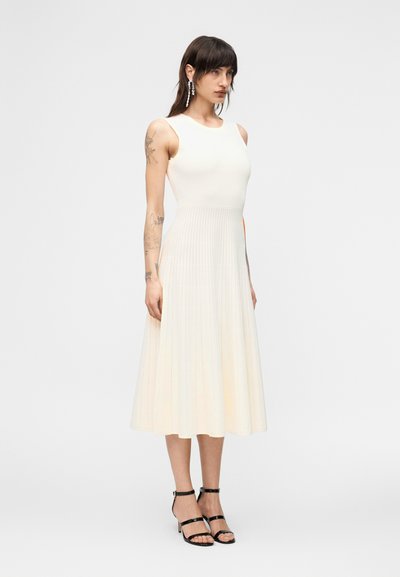 Γυναίκα με τατουάζ που φοράει αμάνικο, κρεμ πλισέ μίντι φόρεμα και μαύρα ψηλοτάκουνα πέδιλα με λουράκια, στέκεται μπροστά σε λευκό φόντο.