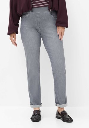 Persona che indossa jeans grigi con vita elasticizzata e orli risvoltati, mocassini neri e giacca color bordeaux sopra una maglia a righe, in piedi davanti a uno sfondo bianco.