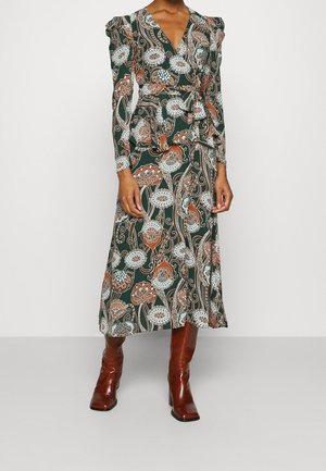 Femme portant une robe longue à manches longues, à motifs floraux, arrivant aux chevilles, avec un col en V et des bottes à talons brunes, sur un fond uni.