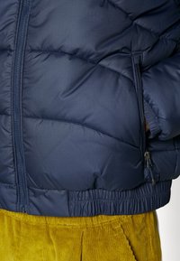 Doudoune marine avec un design matelassé, fermeture éclair sur le devant, taille élastique, accompagnée de pantalons en velours côtelé moutarde visibles en bas.