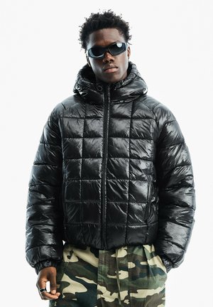 Junger Mann trägt eine schwarze gesteppte Daunenjacke, schwarze Sonnenbrille und Tarnhose, steht mit den Händen in den Taschen frontal da.
