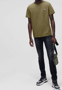 Olivgrünes Baumwoll-T-Shirt mit rundem Ausschnitt und kleinem weißem Logo, kombiniert mit dunklen Jeans und schwarz-weißen Turnschuhen. Kleine Tasche getragen.