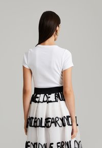 T-shirt blanc à manches courtes associé à une jupe blanche transparente arborant des motifs de texte noirs et une taille noire unie.