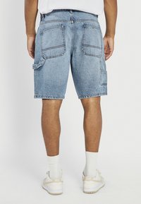 Seljaplaan inimesest, kes kannab helerohelisi teksastest kargo lühkareid, valgeid sokke ja beeži-valgeid Nike tenniseid, seisab ühevärvilise tausta ees.