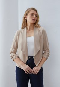 Hellebeige lett blazer med sjalkrage, trekvartlange ermer og brettede mansjetter, båret over en hvit topp og mørkeblå høyt midje jeans.