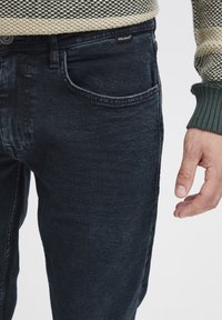 Tamno plave traperice s blago izblijedjelom teksturom, višestrukim džepovima i zatvaranjem na dugme, uparene s puloverom od pletenog uzorka.