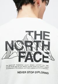 Άσπρο μπλουζάκι με μεγάλο μαύρο γραφικό βουνών και το κείμενο "THE NORTH FACE", μαζί με ένα σλόγκαν στο κάτω μέρος σε έντονη τυπογραφία.