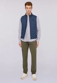 Námořnická prošívaná vesta přes světle šedou košili na zapínání, doplněná olivově zelenými kalhotami a bílými teniskami. Vesta má vodorovné prošívání.