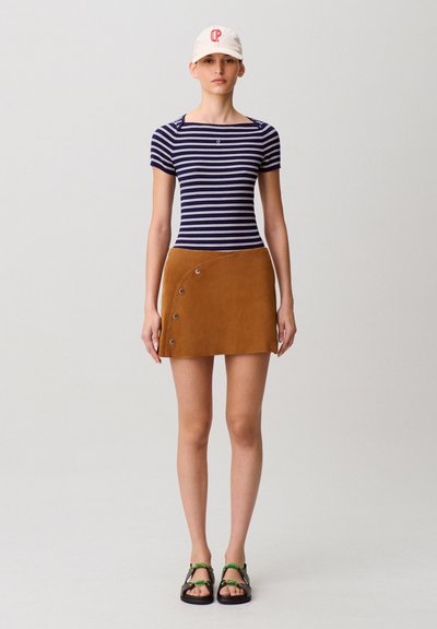 Top a maniche corte a righe blu navy e bianche, minigonna in suede marrone con dettagli a bottoni, cappellino bianco con logo rosso e sandali neri con accenti verdi.