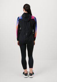 Fekete sportos dzseki kapucnival; színes gradiensekkel díszített ujjak. Fekete leggings-szel és sportcipővel viselve.