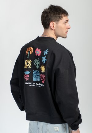 Man in black sweatshirt with colorful abstract floral prints and "Living in Paradise Kaotiko Society" text on the back, wearing light blue jeans.