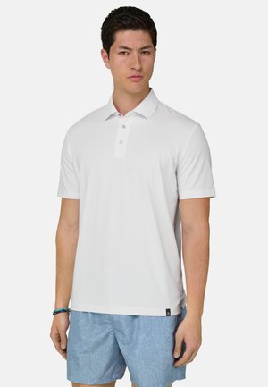 Mann trägt ein weißes kurzärmeliges Poloshirt und hellblaue gemusterte Shorts, steht vor einem schlichten weißen Hintergrund.