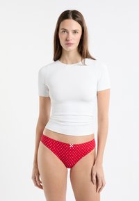 Weißes Kurzarmshirt kombiniert mit roten Bikinihosen mit weißen Punkten und einer weißen Schleife an der Taille, glatter Textur und figurbetontem Design.