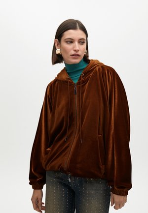 Felpa zip-up in velluto marrone con cappuccio, polsini elasticizzati e due tasche frontali. Indossata sopra un dolcevita in turchese e abbinata a jeans scuri.