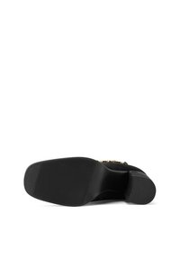 Scarpa nera in suede con tacco alto, suola in gomma texture, punta affusolata e dettagli in metallo dorato sulla parte superiore.