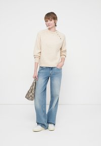 Maglione beige lavorato a maglia con orlo a coste e bottoni decorativi su una spalla, abbinato a jeans a zampa d'elefante blu chiaro e una borsa a fantasia.