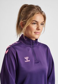 Veste de sport zippée violette avec un col haut, arborant un logo rose clair et des motifs en chevrons sur les épaules. Tissu lisse et extensible.