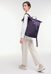 Zaino roll-top viola con spallacci regolabili, dotato di tasca frontale con zip e una texture liscia e opaca. Il modello indossa pantaloni beige larghi.