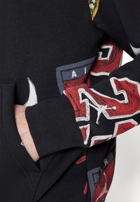 Sudadera negra con diseños gráficos en rojo y gris, con un logo de Jordan. Material de algodón texturizado con bolsillo tipo canguro y puños acanalados.