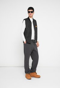 Zwart doorgestikt vest, wit shirt met lange mouwen, grijze losse broek, bruine veterlaarzen en donkere zonnebril, poserend tegen een witte achtergrond.