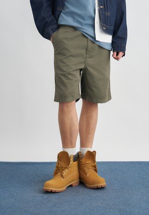 Mand iført grønne shorts, hvide sokker og tan arbejdsstøvler, med den ene hånd i lommen, stående på blåt tæppe mod en ensfarvet baggrund.