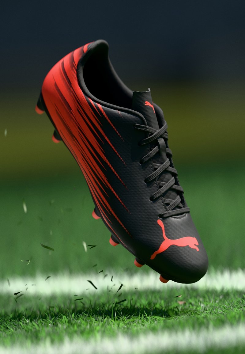 Μαύρο και κόκκινο ποδοσφαιρικό παπούτσι Puma με αγκράφες που σηκώνει γρασίδι και χώμα σε αθλητικό γήπεδο κοντά σε μια λευκή γραμμή.