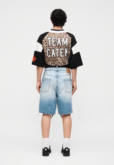 Άτομο με σγουρά μαλλιά που φοράει μαύρο και λεοπάρ φανέλα "TEAM CATEN", ανοιχτό μπλε τζιν σορτς, λευκές κάλτσες και μαύρα αθλητικά παπούτσια με λογότυπο φύλλου σφενδάμου.