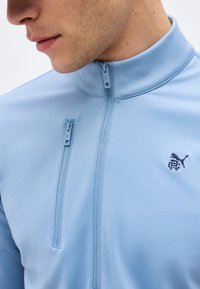 Κοντινό πλάνο ενός ατόμου που φορά ένα γαλάζιο μπουφάν με φερμουάρ και όρθιο γιακά, με μικρό κεντημένο λογότυπο Puma στο στήθος.