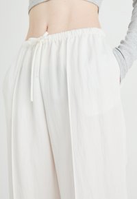 White, lightweight trousers with an elastic waistband and drawstring. Features side pockets and a smooth, flowing fabric texture.