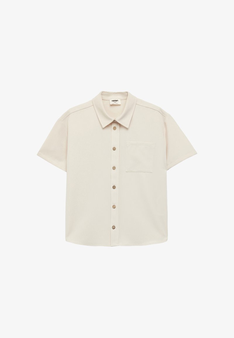 Camisa de manga corta con botones en tejido beige claro. Presenta un cuello apuntado, cinco botones a juego y un bolsillo en el pecho. Textura suave.