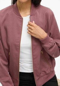 Frau, die eine staubige rosa Bomberjacke über einem weißen Shirt und schwarzen Hosen trägt, hält den Reißverschluss der Jacke nahe am Kragen.
