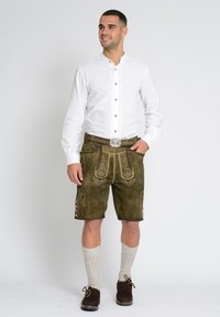 Groene lederhosen met geborduurde details, gecombineerd met een witte blouse met lange mouwen, knielange sokken en donkere schoenen. Zilveren riemgesp als accent.