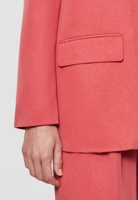 Gros plan d'une main à côté d'une veste de costume rose corail avec une poche à rabat et un pantalon assorti.